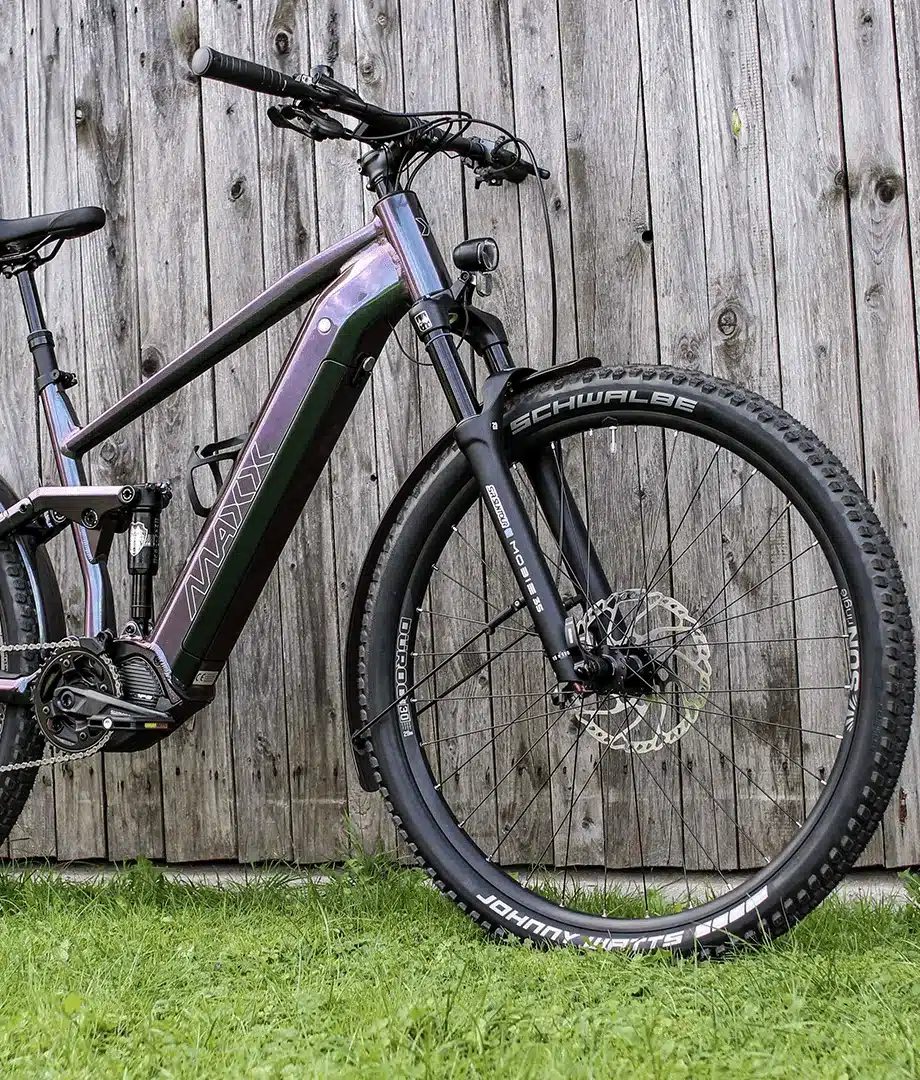 Seitenansicht eines MAXX Tourmaxx ELS Tour E-Trekking-SUV mit Vollfederung, das an eine Holzplankenwand gelehnt ist.
