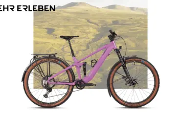 Lila MAXX Jinxx ELF Tour E-Bike mit Vollfederung, Vario-Sattelstütze, Gepäckträger, Schutzblechen und integrierter Lichtanlage, abgebildet vor einer Kulisse von Hügeln.