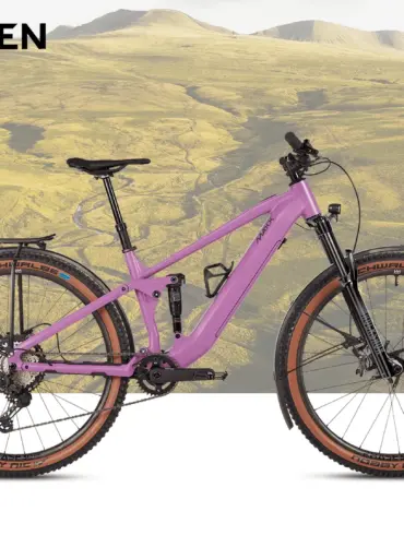 Lila MAXX Jinxx ELF Tour E-Bike mit Vollfederung, Vario-Sattelstütze, Gepäckträger, Schutzblechen und integrierter Lichtanlage, abgebildet vor einer Kulisse von Hügeln.