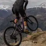 Ein Mountainbiker fährt mit einem lila E-Mountainbike auf einem felsigen Bergpfad mit schneebedeckten Gipfeln im Hintergrund.