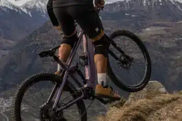 Ein Mountainbiker fährt mit einem lila E-Mountainbike auf einem felsigen Bergpfad mit schneebedeckten Gipfeln im Hintergrund.