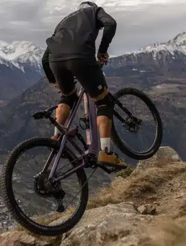 Ein Mountainbiker fährt mit einem lila E-Mountainbike auf einem felsigen Bergpfad mit schneebedeckten Gipfeln im Hintergrund.