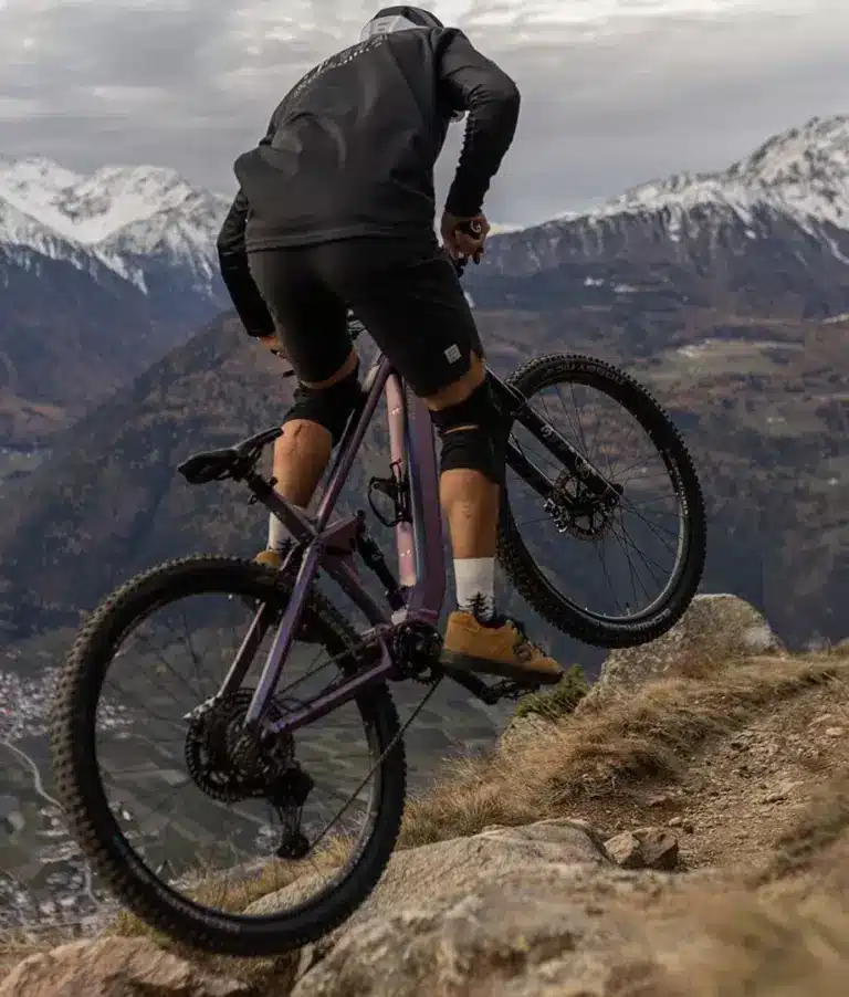 Ein Mountainbiker fährt mit einem lila E-Mountainbike auf einem felsigen Bergpfad mit schneebedeckten Gipfeln im Hintergrund.
