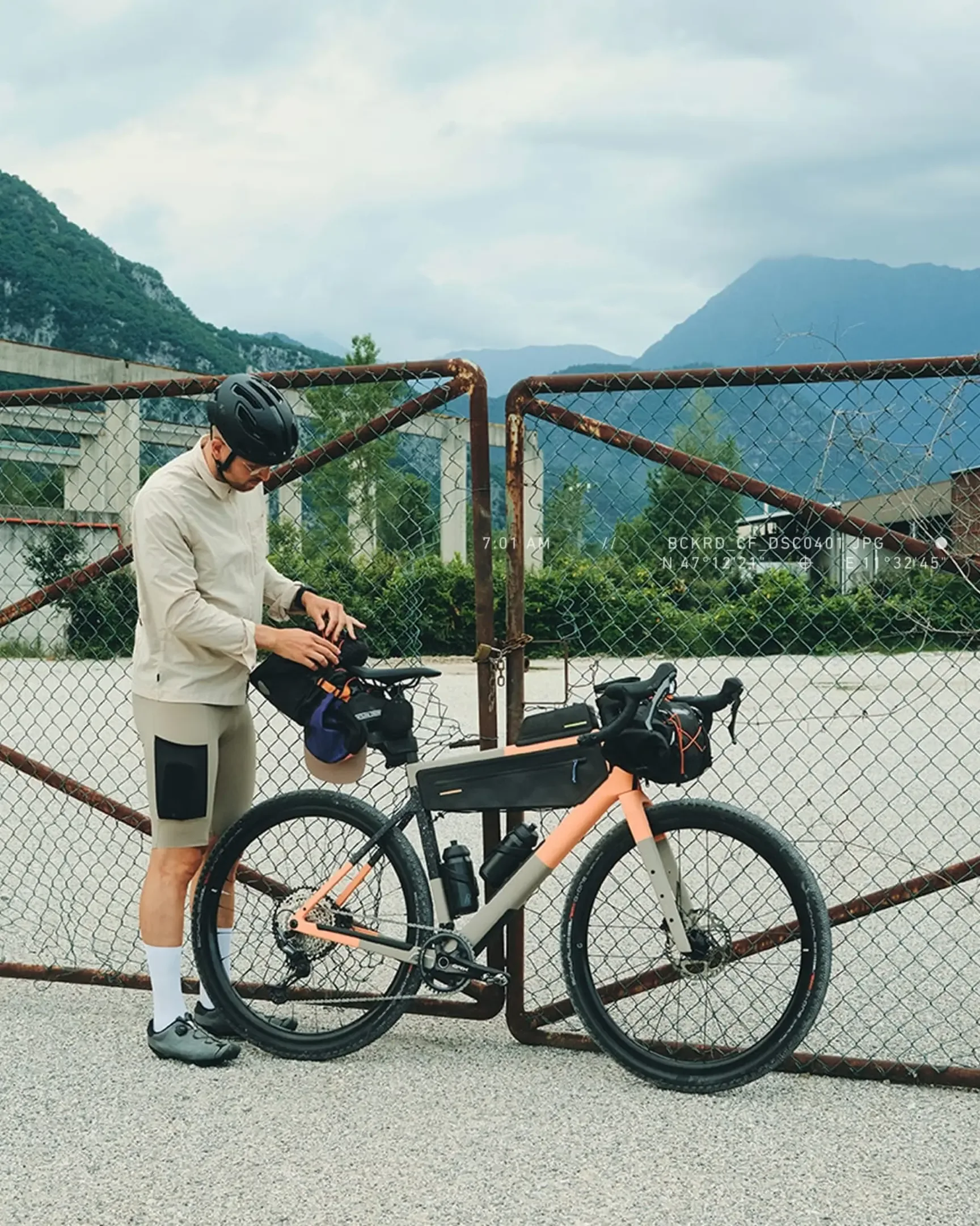 Mann im Gravel-Outfit überprüft die Anbringung von Bikepacking-Taschen an einem Backroad Gravelbike vor einem Metallzaun.