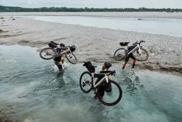 Drei Radfahrer tragen ihre mit Gepäcktaschen versehenen Gravelbikes durch flaches Wasser eines Kiesstrands, im Hintergrund ein breiter Fluss und Bäume.