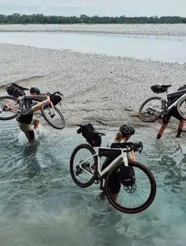 Drei Radfahrer tragen ihre mit Gepäcktaschen versehenen Gravelbikes durch flaches Wasser eines Kiesstrands, im Hintergrund ein breiter Fluss und Bäume.