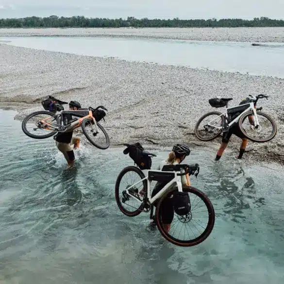 Drei Radfahrer tragen ihre mit Gepäcktaschen versehenen Gravelbikes durch flaches Wasser eines Kiesstrands, im Hintergrund ein breiter Fluss und Bäume.