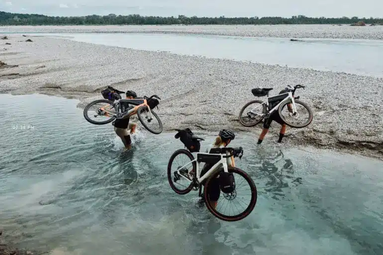 Drei Radfahrer tragen ihre mit Gepäcktaschen versehenen Gravelbikes durch flaches Wasser eines Kiesstrands, im Hintergrund ein breiter Fluss und Bäume.