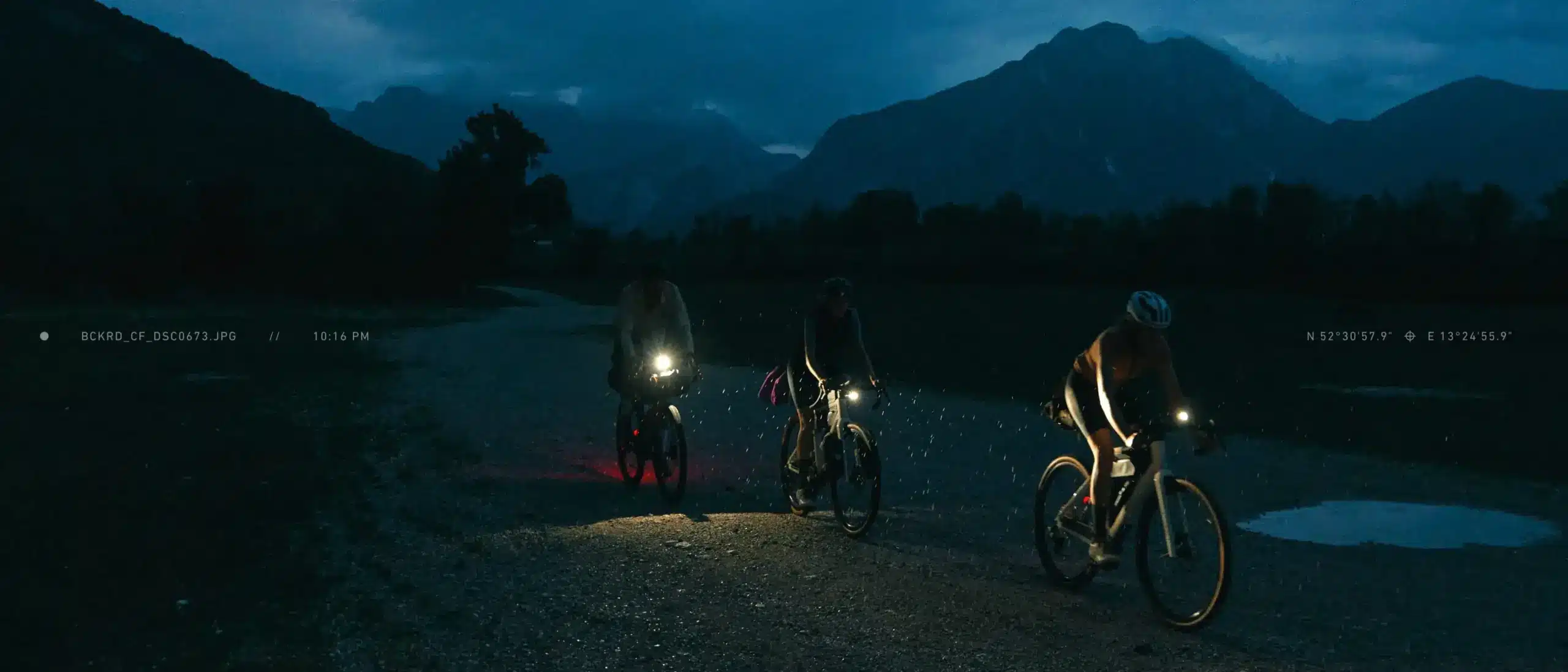 Drei Personen fahren nachts mit Gravelbikes auf einem Schotterweg in bergiger Landschaft, beleuchtet von ihren Vorder- und Rücklichtern.
