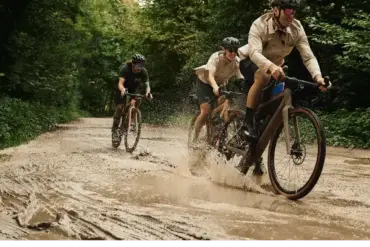 Drei Personen fahren mit Gravelbikes auf einer matschigen Straße durch einen grünen Wald, wobei das vordere Fahrrad Wasser aufwirbelt.