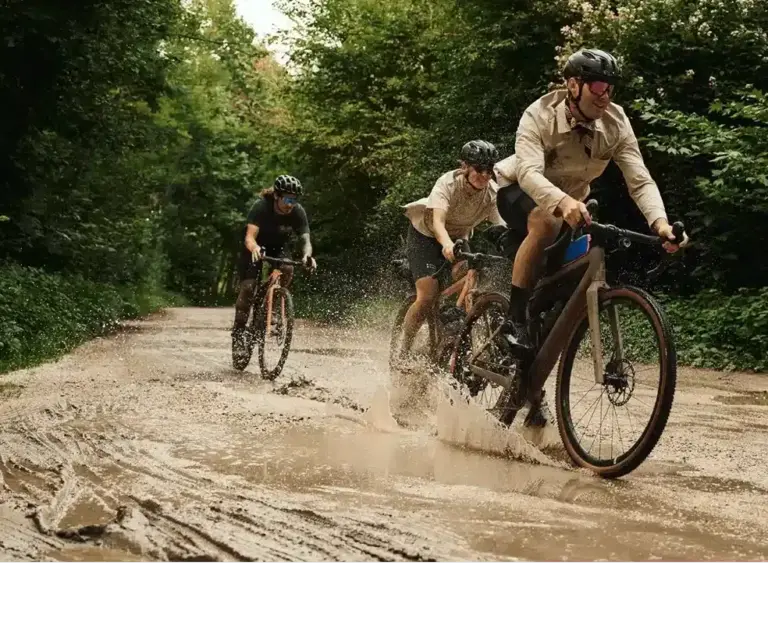 Drei Personen fahren mit Gravelbikes auf einer matschigen Straße durch einen grünen Wald, wobei das vordere Fahrrad Wasser aufwirbelt.