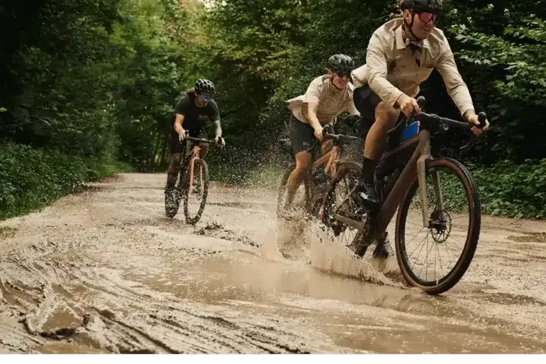 Drei Personen fahren mit Gravelbikes auf einer matschigen Straße durch einen grünen Wald, wobei das vordere Fahrrad Wasser aufwirbelt.