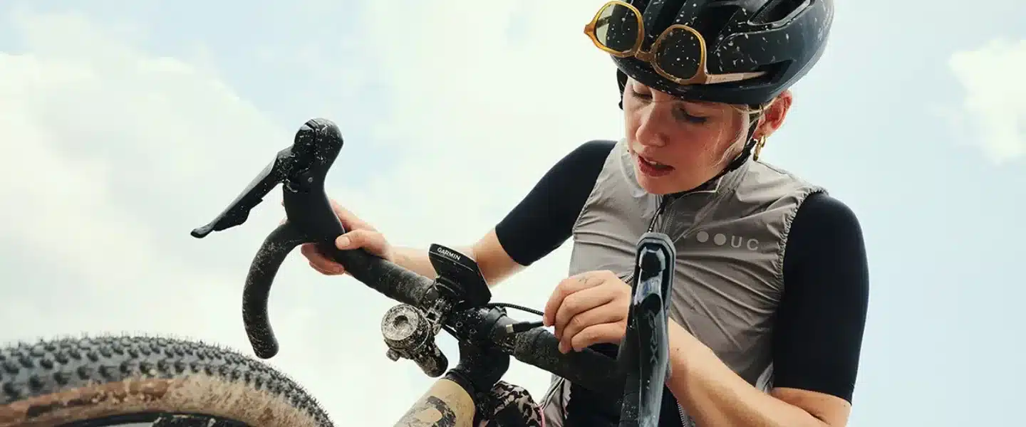 Radsportlerin, die ihr Gravelbike für eine Tour vorbereitet. Sie trägt einen Helm und eine Weste. Das Fahrrad ist schlammig, was auf eine Geländefahrt hindeutet.