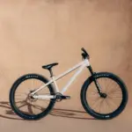 Ein weißes Rose Bruce 1 Dirt Jump Bike mit schwarzen Maxxis-Reifen, Gabel und Sattel steht seitlich vor einer sandfarbenen Wand.
