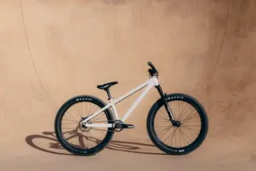 Ein weißes Rose Bruce 1 Dirt Jump Bike mit schwarzen Maxxis-Reifen, Gabel und Sattel steht seitlich vor einer sandfarbenen Wand.