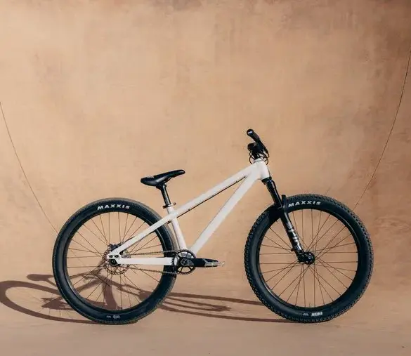 Ein weißes Rose Bruce 1 Dirt Jump Bike mit schwarzen Maxxis-Reifen, Gabel und Sattel steht seitlich vor einer sandfarbenen Wand.