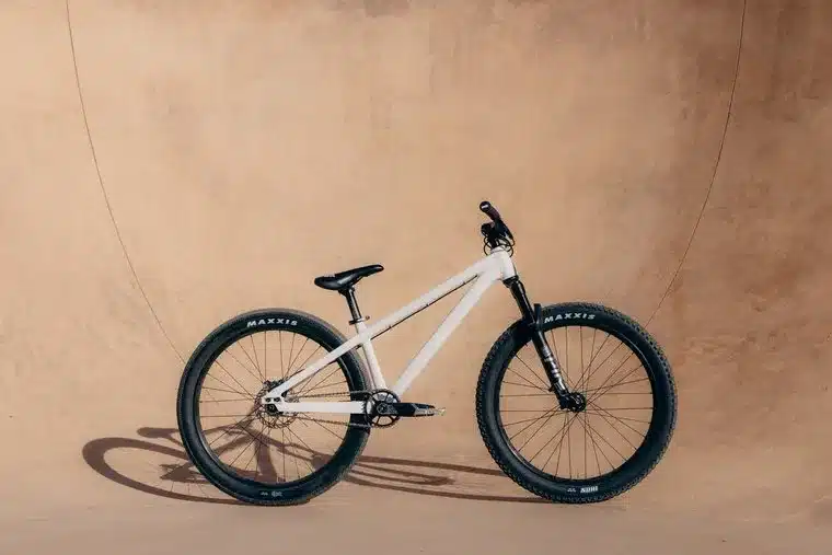 Ein weißes Rose Bruce 1 Dirt Jump Bike mit schwarzen Maxxis-Reifen, Gabel und Sattel steht seitlich vor einer sandfarbenen Wand.