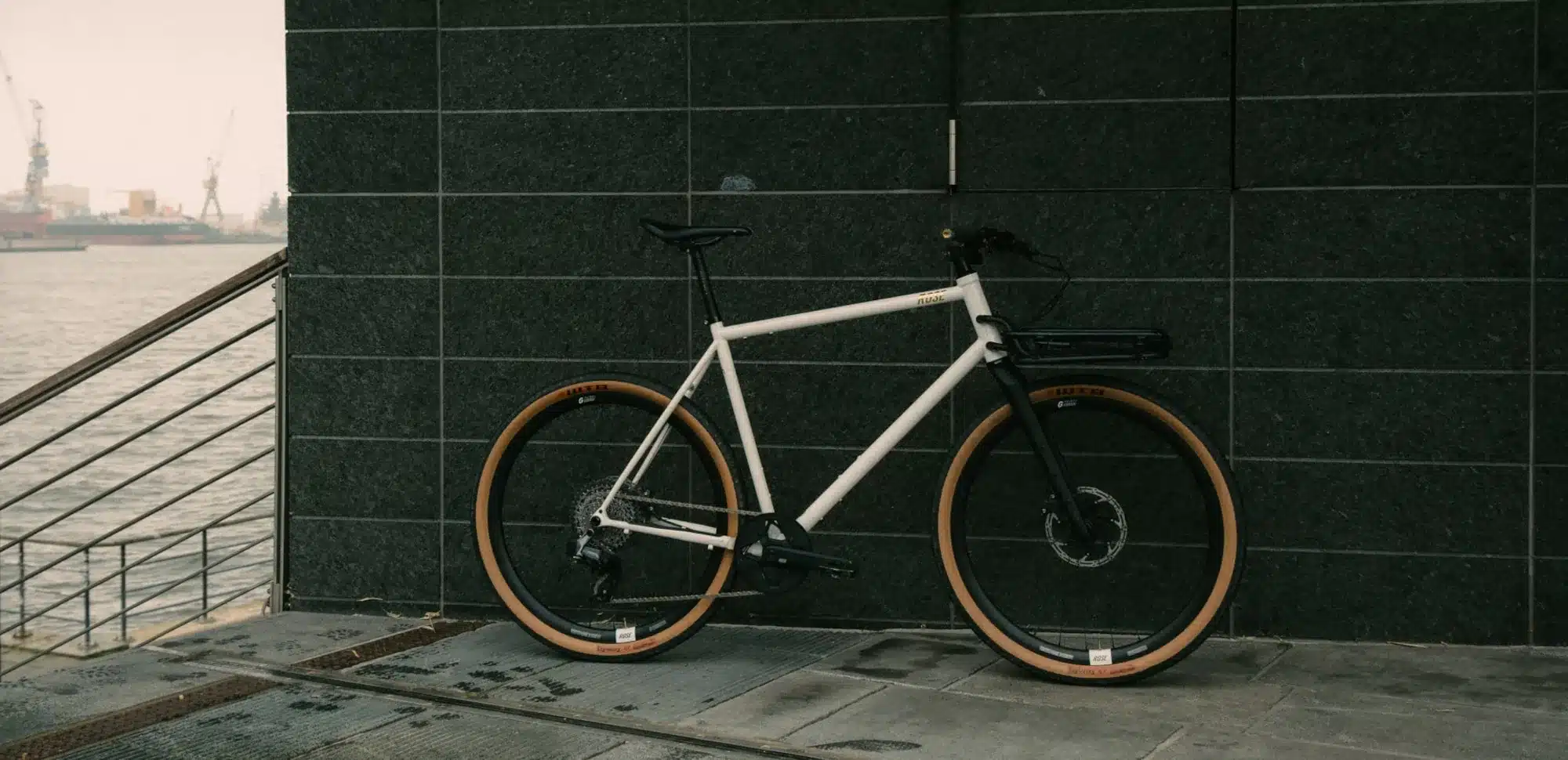 Ein weißes ROSE Hobo Gravelbike mit schwarzem Frontträger und braunen Reifen steht vor einer dunklen, gefliesten Wand, mit Blick auf einen Hafen im Hintergrund.