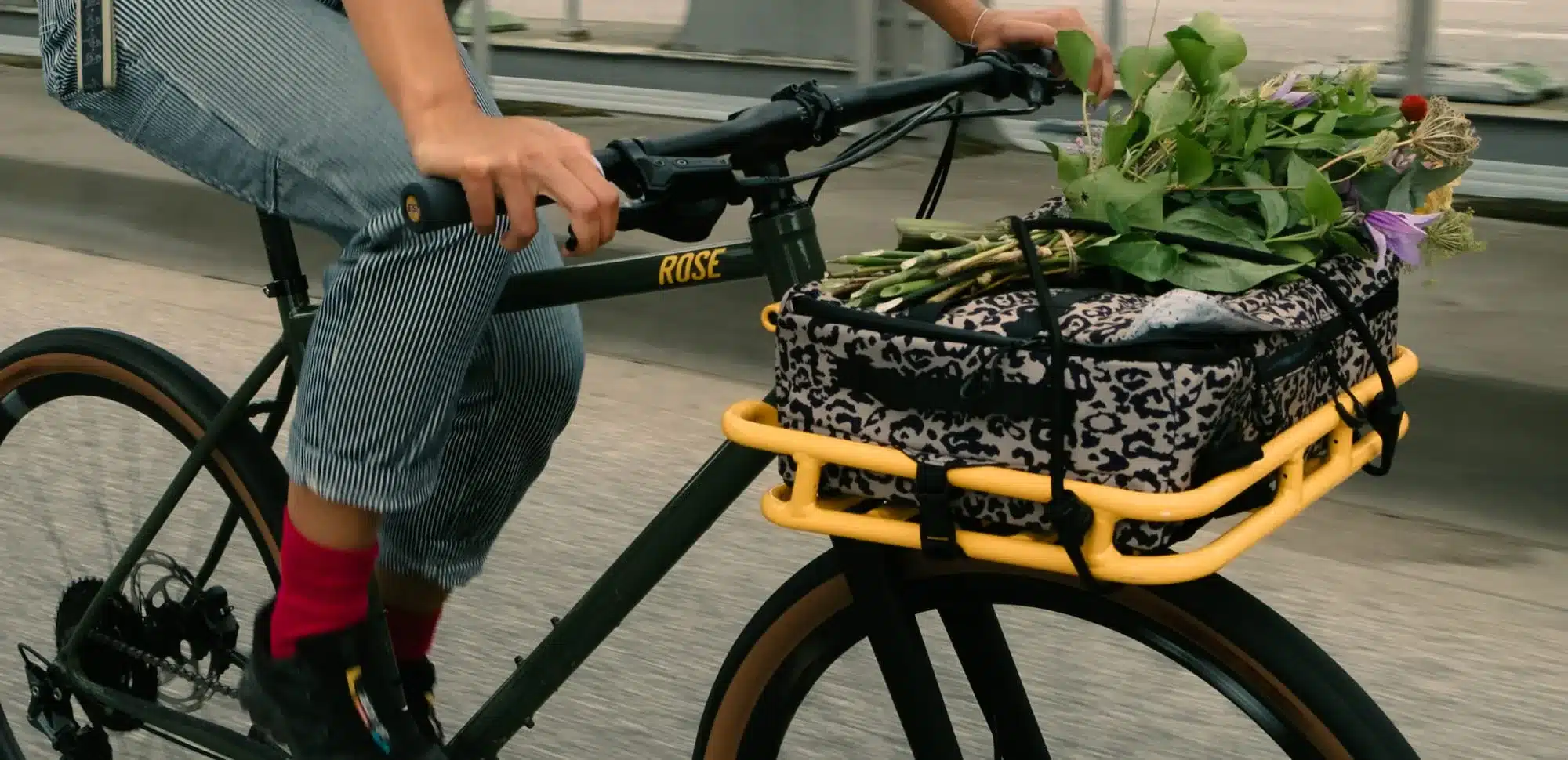 Eine Person fährt ein dunkles Rose Hobo Fahrrad mit einem auffälligen gelben Frontträger, der mit einer Leo-Muster-Tasche und frischen Blumen beladen ist.