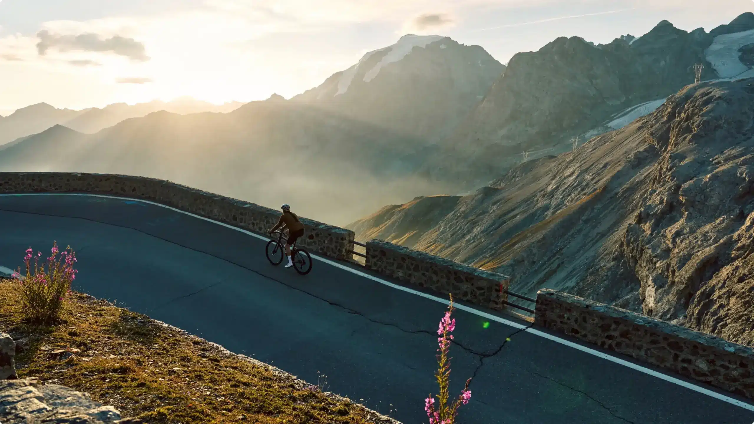 Ein Rennradfahrer fährt auf einer asphaltierten Straße mit Steinbegrenzung einen Bergpass hinauf. Im Hintergrund erheben sich schroffe Berge und Gletscher im warmen Licht der untergehenden Sonne.