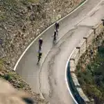Drei Rennradfahrer fahren bergauf auf einer kurvenreichen Straße mit Steinmauern auf beiden Seiten.