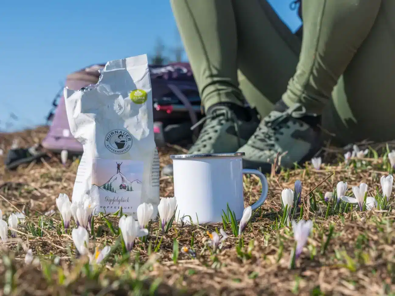 Kaffeepackung „Gipfelglück“ der Murnauer Kaffeerösterei und Emaillebecher beim Wandern auf einer Wiese mit Krokussen.