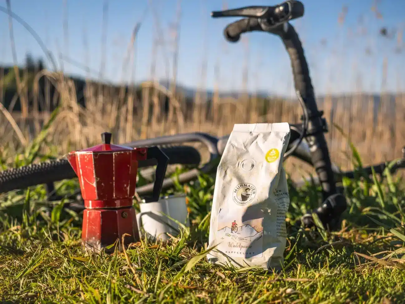 Packung Murnauer Kaffeerösterei 'Wadelpause' neben Espressokocher und Fahrradlenker auf einer Wiese.