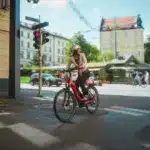 Eine Frau mit Helm fährt ein rotes Winora Sinus R5f E-Bike durch eine Stadtstraße, während sie auf eine rote Ampel wartet.