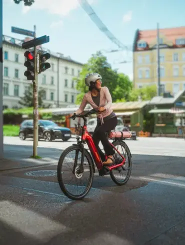 Eine Frau mit Helm fährt ein rotes Winora Sinus R5f E-Bike durch eine Stadtstraße, während sie auf eine rote Ampel wartet.