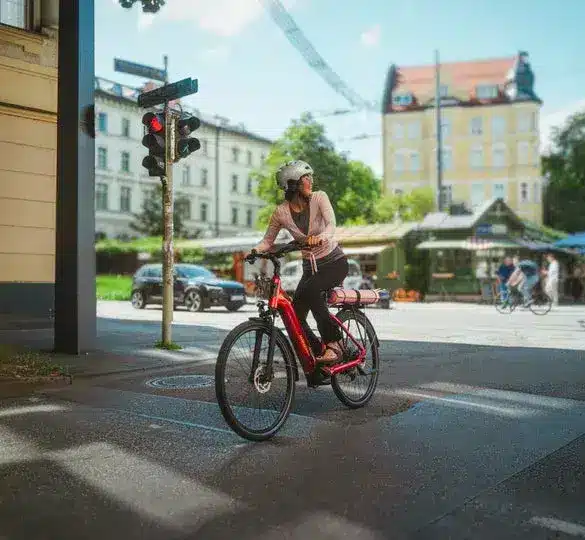 Eine Frau mit Helm fährt ein rotes Winora Sinus R5f E-Bike durch eine Stadtstraße, während sie auf eine rote Ampel wartet.