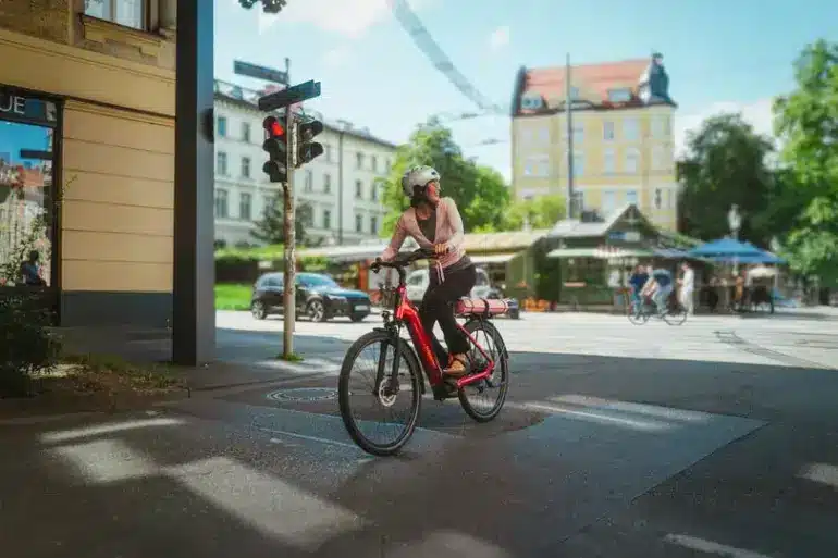 Eine Frau mit Helm fährt ein rotes Winora Sinus R5f E-Bike durch eine Stadtstraße, während sie auf eine rote Ampel wartet.