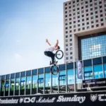 BMX-Fahrer in der Luft mit seinem Fahrrad, eine moderne Stadtkulisse im Hintergrund.