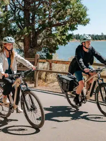Mann und Frau fahren nebeneinander auf Fiido Nomads E-Bikes auf einem asphaltierten Weg neben einem See.