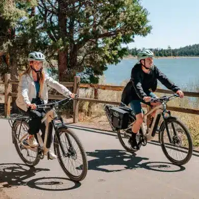 Mann und Frau fahren nebeneinander auf Fiido Nomads E-Bikes auf einem asphaltierten Weg neben einem See.