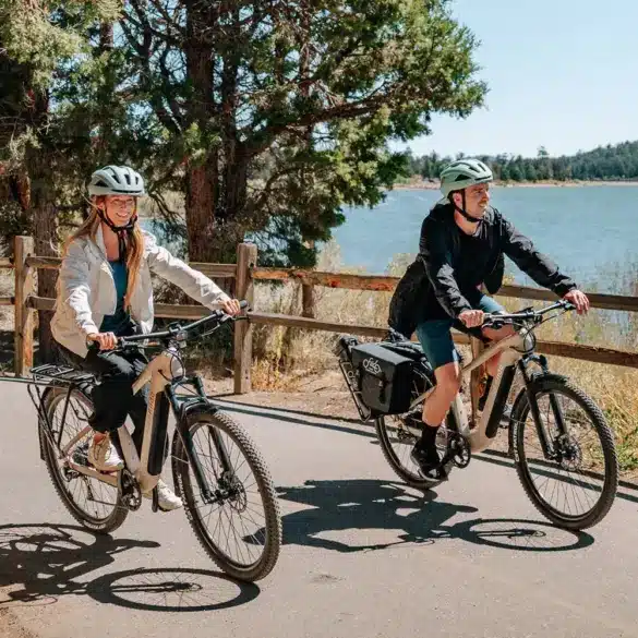 Mann und Frau fahren nebeneinander auf Fiido Nomads E-Bikes auf einem asphaltierten Weg neben einem See.