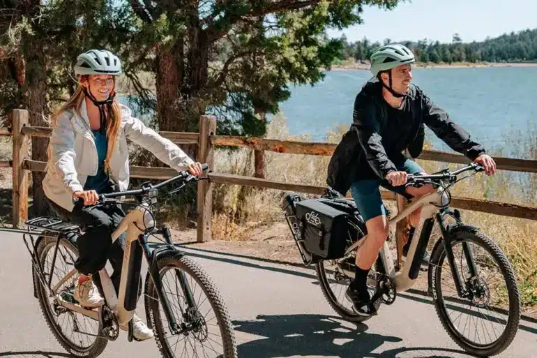 Mann und Frau fahren nebeneinander auf Fiido Nomads E-Bikes auf einem asphaltierten Weg neben einem See.