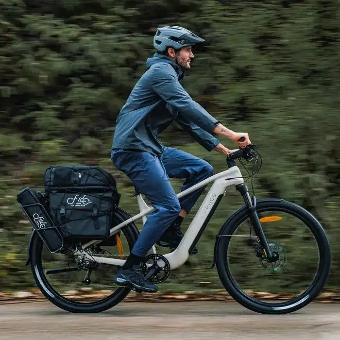 Ein Mann in dunkler Kleidung und Helm fährt ein weißes Fiido Nomads E-Bike mit vollen Gepäcktaschen auf dem Rücksitz auf einer asphaltierten Straße.