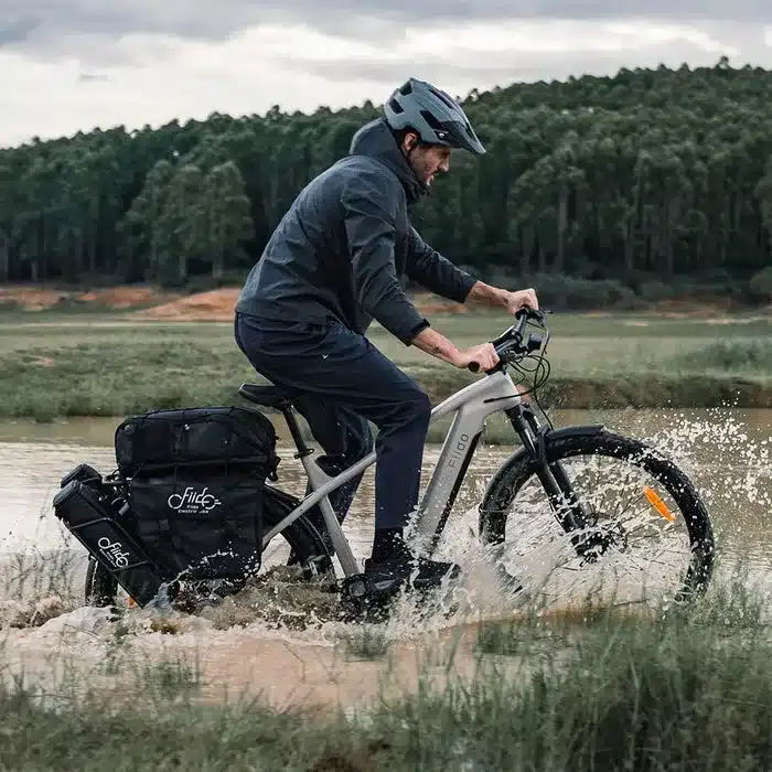 Mann mit Helm fährt Fiido Nomads E-Bike durch aufspritzendes Wasser in einer ländlichen Umgebung.