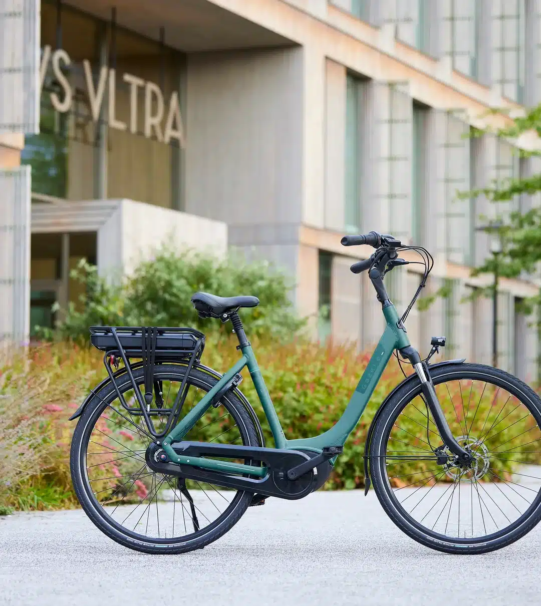 Seitenansicht eines grünen Gazelle Paris E-Bikes mit Gepäckträger, Akku und Bosch-Mittelmotor, geparkt auf einem Gehweg vor einem modernen Gebäude.
