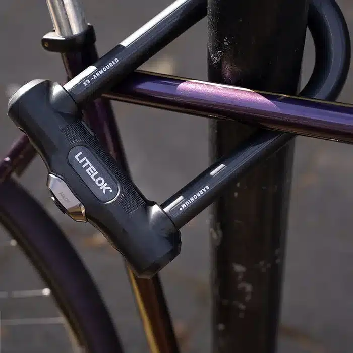 Ein LITELOK X3 Bügelschloss sichert ein Fahrrad an einem dunklen Pfosten. Das Schloss ist schwarz und hat auf dem Bügel den Schriftzug Barronium. Das Fahrrad hat einen violetten Rahmen.