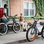 Zwei Ruff Cycles Biggie E-Bikes, eines in Schwarz mit weißem Reifengummi und eines in Blau mit orangen Felgen, stehen auf einem Gehweg vor einem Backsteinhaus. Ein Mann mit einem roten Rucksack auf dem Rücken und eine Frau mit einer Trinkflasche auf einer Sitzbank sind im Bild.