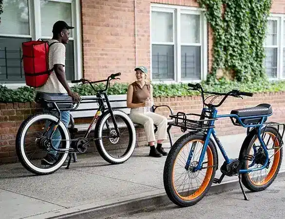 Zwei Ruff Cycles Biggie E-Bikes, eines in Schwarz mit weißem Reifengummi und eines in Blau mit orangen Felgen, stehen auf einem Gehweg vor einem Backsteinhaus. Ein Mann mit einem roten Rucksack auf dem Rücken und eine Frau mit einer Trinkflasche auf einer Sitzbank sind im Bild.