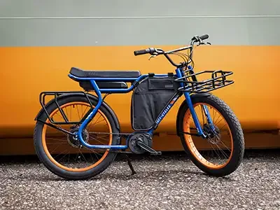 Ein blaues Ruff Cycles Biggie E-Bike mit orangefarbenen Felgen, schwarzem Gepäckträger vorne und hinten, einer schwarzen Tasche am Rahmen und BMX-Lenker, abgestellt auf Schotter vor einem orange-grauen Hintergrund.