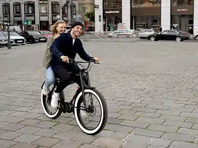 Mann und Frau fahren gemeinsam auf einem schwarzen Ruff Cycles Biggie E-Bike mit breiten weißen Reifen auf einem gepflasterten Platz.