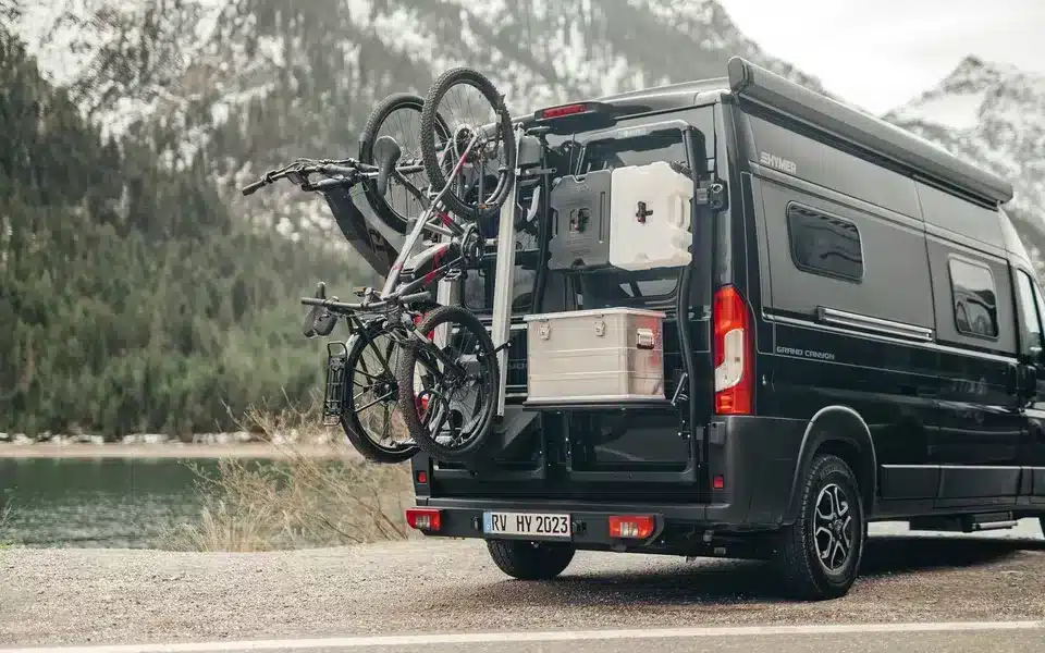 Bike Carrier Swing Fahrradträger mit zwei Fahrrädern am Heck eines Vans, daneben Alukoffer und Campingausrüstung, im Hintergrund Berge und See.