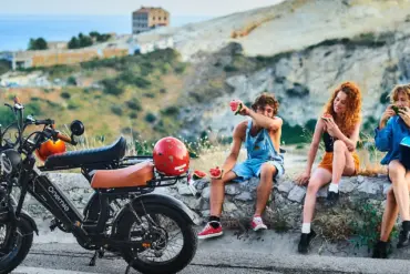 Ein Onemile Scrambler E-Bike steht neben einer Gruppe junger Leute, die auf einer Steinmauer sitzen und Wassermelone essen.