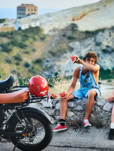 Ein Onemile Scrambler E-Bike steht neben einer Gruppe junger Leute, die auf einer Steinmauer sitzen und Wassermelone essen.