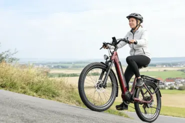 Frau mit Helm auf einem roten Dukawey FUGL3.0 City-E-Bike fährt auf einer Straße bergauf.