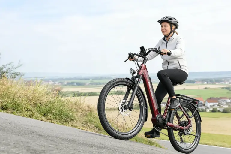 Frau mit Helm auf einem roten Dukawey FUGL3.0 City-E-Bike fährt auf einer Straße bergauf.
