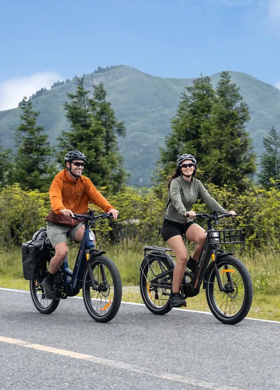 Ein Mann in orangefarbener Jacke und eine Frau in olivgrüner Jacke fahren nebeneinander auf ENGWE E26 3.0 Pro E-Bikes auf einer asphaltierten Straße.